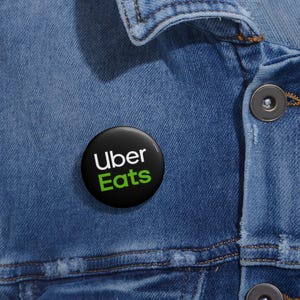 May include: A black button featuring the Uber Eats logo in white and green. The button is attached to a blue denim jacket. The button is circular and has a shiny surface.
