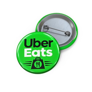 Pode incluir: Um broche redondo com alfinete, apresentando um fundo verde brilhante, o texto Uber Eats em preto e branco e um gráfico preto de uma sacola de comida com um ícone de garfo e faca. A parte de trás mostra um mecanismo de metal prateado.