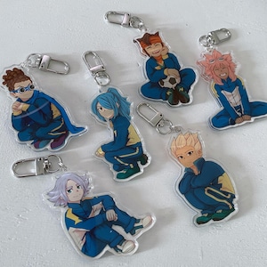 Könnte beinhalten: Sechs bunte Anime-Charakter-Schlüsselanhänger mit blauen, gelben, orangen, rosa und weißen Akzenten. Die Schlüsselanhänger bestehen aus klarem Acryl und zeigen verschiedene Charaktere aus der Anime-Serie Inazuma Eleven.