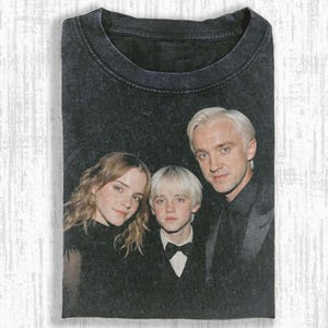 May include: Dark gray t-shirt featuring a portrait of three people. The image shows a woman with long wavy hair, a young person with short, light-colored hair, and a man with short, light-colored hair. All are dressed in dark clothing.
