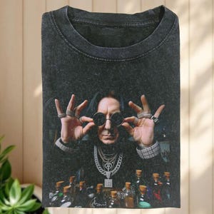 May include: Dark gray t-shirt featuring a portrait of a man wearing round sunglasses and multiple rings. The man is also wearing several necklaces and bracelets. Below the portrait are several bottles with cork stoppers.
