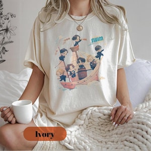 May include: Ivory-colored t-shirt featuring a cartoon illustration of a ship with several animated characters. The word "SWIM" is above the ship. The shirt is a neutral color and has short sleeves.
