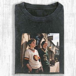 May include: Dark gray t-shirt featuring a graphic of two people standing outside a building. One person wears a cream-colored shirt with a lion crest, the other a green sweater with a snake. The image is from a movie.