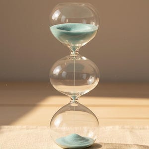 May include: A clear glass hourglass with light blue sand. The hourglass has a bulbous shape with three rounded sections. The sand is flowing from the top section to the bottom section. The hourglass is sitting on a beige surface.