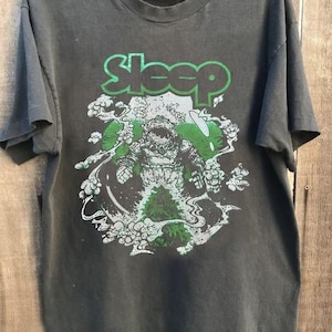 Könnte beinhalten: Ein schwarzes Kurzarm-T-Shirt mit einem Grafikdruck eines Astronauten, der von grünem Rauch und Wolken umgeben ist. Das Wort Sleep ist in großen grünen Buchstaben über dem Astronauten gedruckt. Das T-Shirt hat einen Rundhalsausschnitt.