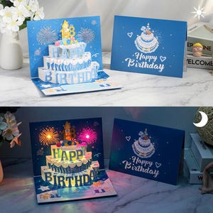 Può includere: Biglietto di compleanno pop-up blu con una torta a tre piani e la scritta "Happy Birthday". Il biglietto presenta un disegno di fuochi d'artificio e un biglietto più piccolo con un'illustrazione di torta. Il biglietto pop-up si illumina.