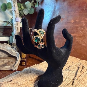 May include: A black velvet hand-shaped jewelry display holding three rings. The rings feature green gemstones and intricate wire wrapping in gold and copper tones. The display sits on a wooden surface, showcasing the rings' details.
