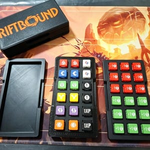Pode incluir: Uma caixa de jogo preta com a palavra "RIFTBOUND" em letras laranja. Ao lado, duas bandejas pretas com peças de jogo coloridas. As peças de jogo são vermelhas, verdes, azuis, amarelas e roxas, com símbolos e números.