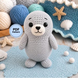 May include: A gray crocheted seal plush toy with a cream-colored muzzle, black safety eyes, and a black stitched nose and mouth. A circular blue badge with the text PDF Pattern is visible. The seal is surrounded by yarn balls and seashells.