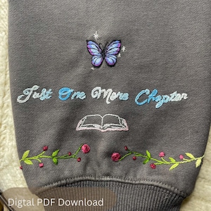 May include: Dark gray sweatshirt with embroidered butterfly, text, and floral design. The butterfly is purple and blue. The text reads "Just One More Chapter" in blue and white. A pink book and green and red floral design are also embroidered.