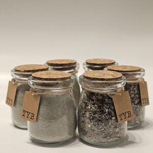 May include: Six small glass jars with cork lids, filled with various granular substances. Each jar has a small brown tag with the letters "TYB" tied around the neck. The jars are arranged in a circular pattern.