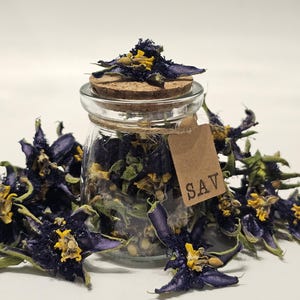 May include: A clear glass jar with a cork stopper filled with dried dark blue and yellow flowers. A small brown paper tag tied with twine around the neck of the jar features the letters SAV printed in black. More dried flowers surround the base.