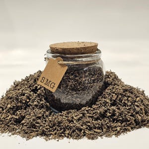 May include: A clear glass jar filled with dried herbs, sealed with a cork stopper, and a small tag that reads "SMG". The jar sits on a bed of the same dried herbs, all against a white background. The herbs are a muted brown color.