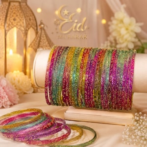 May include: A collection of colorful bangles in shades of green, yellow, pink, and purple, displayed on a white stand. The bangles are adorned with glitter and are arranged in a decorative manner. The background includes a gold lantern and the text "Eid Mubarak".