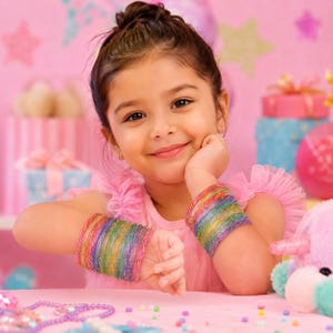 May include: A young girl smiles, wearing a pink dress and two rainbow-colored wrist cuffs. The cuffs are made of colorful threads, with hues of red, orange, yellow, green, blue, and purple. The background is pink with gift boxes and a stuffed animal.