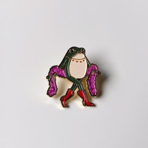 Puede incluir: Un broche de esmalte y metal dorado con forma de rana verde de vientre crema, que lleva botas rojas y un collar fino con pequeñas estrellas rojas. La rana está envuelta en una larga boa de color púrpura texturizada que cuelga de sus brazos.