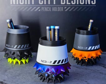 Könnte beinhalten: Drei Stifthalter mit futuristischem Design. Die Halter sind schwarz, weiß und grau mit Neon-Akzenten. Jeder Halter hat eine stachelige Basis und den Text "NCD-2". Der obere Text lautet "NIGHT CITY DESIGNS PENCIL HOLDER".