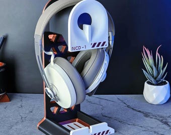 May include: A black, white, and copper headphone stand holding a pair of grey and white headphones. The stand features a geometric design with the text "NCD-1" and diagonal stripes. A small succulent plant in a white pot is visible in the background.