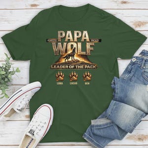 May include: A dark green t-shirt with the words "PAPA WOLF" in gold lettering, featuring a wolf silhouette and the text "LEADER OF THE PACK." Below are paw prints with names. White sneakers and blue jeans are also visible.