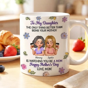 May include: White ceramic mug with a handle, decorated with a graphic of two women holding coffee cups. The mug has text that reads "To My Daughter" and "Happy Mother's Day". Floral and leaf accents surround the text and images.