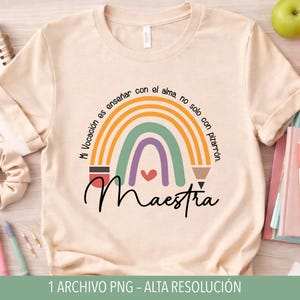 Puede incluir: Camiseta beige con un diseño de arcoíris y la palabra "Maestra" en escritura negra. El arcoíris presenta colores naranja, verde, morado y rojo. El texto alrededor del arcoíris dice "Mi Vocación es enseñar con el alma, no solo con pizarrón."