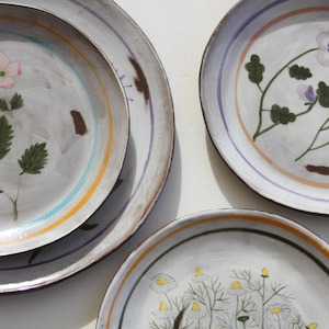 May include: A selection of hand-painted ceramic plates. The plates showcase floral designs in pink, purple, and yellow, with green leaves and stems. Each plate has a grey background with colourful striped borders.