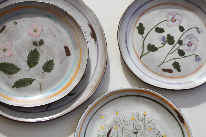 May include: A selection of hand-painted ceramic plates. The plates showcase floral designs in pink, purple, and yellow, with green leaves and stems. Each plate has a grey background with colourful striped borders.