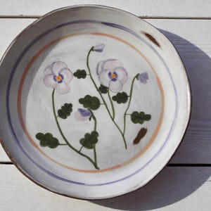 May include: A round, hand-painted ceramic plate with a white base and a floral design. The plate features a light blue and orange border, and the floral design includes purple pansies and green leaves. The plate is approximately 25 cm in diameter.