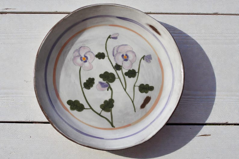May include: A round, hand-painted ceramic plate with a white base and a floral design. The plate features a light blue and orange border, and the floral design includes purple pansies and green leaves. The plate is approximately 25 cm in diameter.