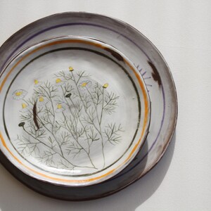 May include: Two ceramic plates, one stacked on the other, with a floral design. The top plate is white with a hand-painted botanical illustration in green, yellow, and brown. The bottom plate is grey with a purple and orange stripe.