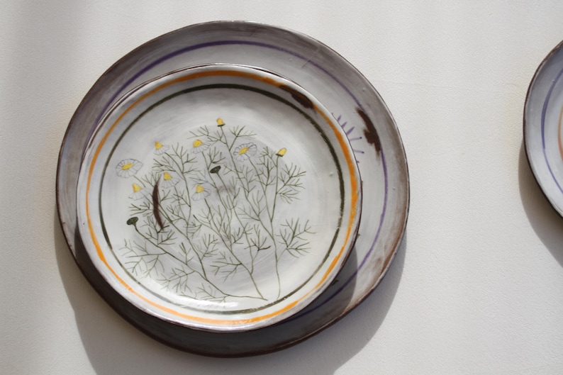 May include: Two ceramic plates, one stacked on the other, with a floral design. The top plate is white with a hand-painted botanical illustration in green, yellow, and brown. The bottom plate is grey with a purple and orange stripe.