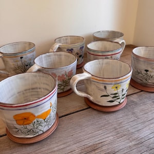 Può includere: Una collezione di tazze in ceramica con superficie smaltata bianco sporco, ognuna caratterizzata da illustrazioni botaniche dipinte a mano di vari fiori selvatici nei toni dell'arancione, giallo e rosa, con basi in terracotta e manici semplici.