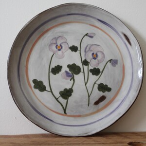 May include: A round ceramic plate with a hand-painted floral design. The plate has a light grey background with a purple and orange border. The flowers are lavender with green leaves.