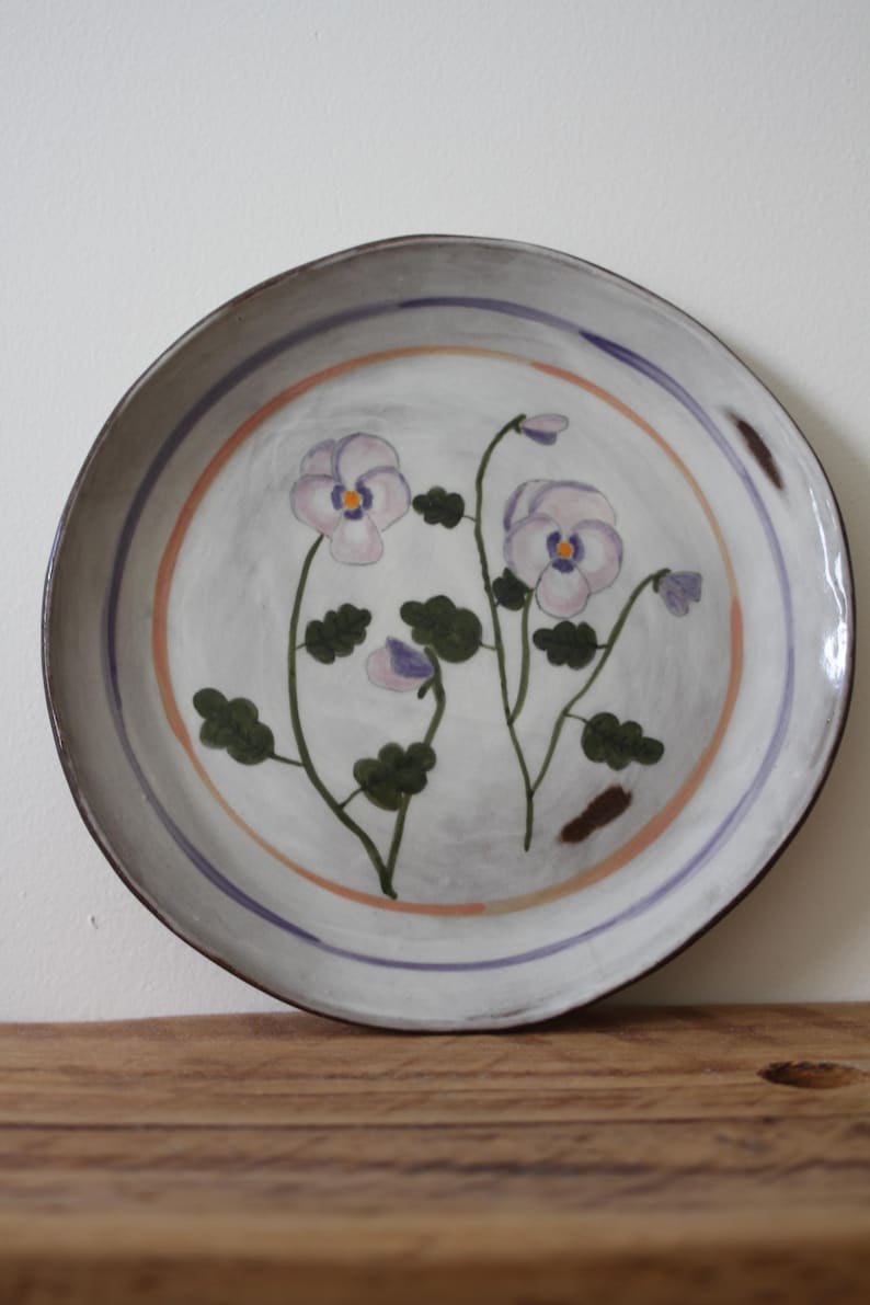 May include: A round ceramic plate with a hand-painted floral design. The plate has a light grey background with a purple and orange border. The flowers are lavender with green leaves.