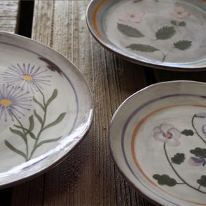 May include: Three oval ceramic plates with floral designs. Each plate features a different flower: purple daisies, pink flowers, and purple pansies. The plates have a grey background with a brown rim and colourful accent stripes.