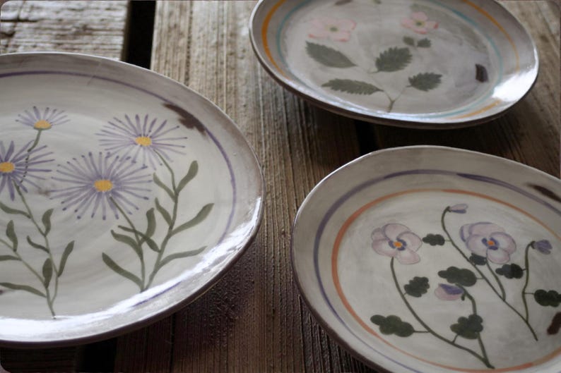 May include: Three oval ceramic plates with floral designs. Each plate features a different flower: purple daisies, pink flowers, and purple pansies. The plates have a grey background with a brown rim and colourful accent stripes.