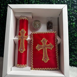 May include: A red religious gift set in a white box. The set includes a candle, a book, a small silver container, and a small bag. The candle and book feature gold crosses and trim. The items are arranged against a green background.