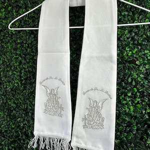 May include: White clergy stole with silver embroidered angel design and the text "Recuerda De Mi Mensaje". The stole has a fringed bottom and is hanging on a white hanger against a green background.