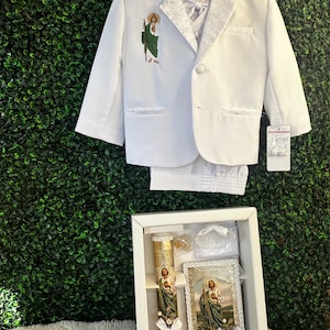 May include: A white suit set for a special occasion, featuring a blazer with embroidered details and matching pants. The set includes a vest and bow tie. A gift box contains a candle, a framed image, and other accessories, all with religious imagery.