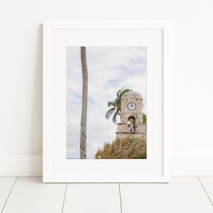 Puede incluir: Impresión enmarcada de una torre del reloj de piedra con una esfera de reloj, palmeras y un cielo nublado. La torre está rodeada de hierba alta. La impresión está en un marco blanco y se muestra en una pared blanca.