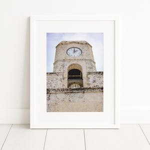 Puede incluir: Impresión enmarcada de una torre del reloj desgastada, de color blanquecino, sobre un cielo azul pálido. La esfera del reloj muestra la hora. La arquitectura incluye aberturas arqueadas y mampostería texturizada, creando una estética vintage.