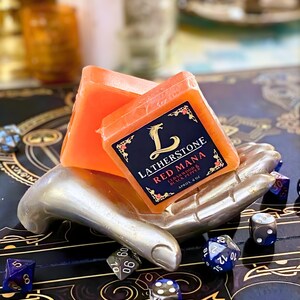 Pode incluir: Duas barras de sabão laranja repousam em um suporte prateado em forma de mão, com dados espalhados ao redor. O sabão é rotulado como "Latherstone Red Mana" com "Cedarwood & Black Pepper" e "Aprox. 113g" impresso no rótulo. Os dados são de várias cores e formas.