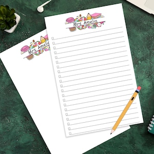 May include: Two white notepads with lined pages and check boxes. The top notepad has the text "Mrs. Amelia" above a colourful illustration of school supplies. A yellow pencil with a pink eraser rests on the bottom notepad.