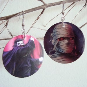 May include: A pair of round earrings with a silver hook. One earring features a black and white illustration of a man in a mask and cape. The other earring features a black and white illustration of a mummy with red eyes.