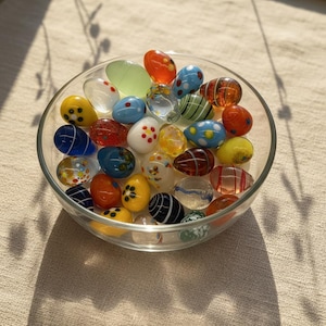 May include: A glass bowl containing a collection of decorative glass eggs. The eggs are in various colours and patterns, including polka dots, stripes, and floral designs. The colours include red, blue, yellow, orange, and green.