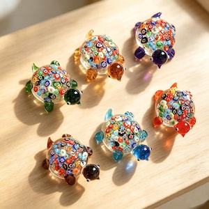 May include: Six glass turtle figurines with clear shells filled with multicolored millefiori floral patterns. Each turtle has a solid colored glass head and four flippers in shades of green, amber, purple, orange, blue, and brown.