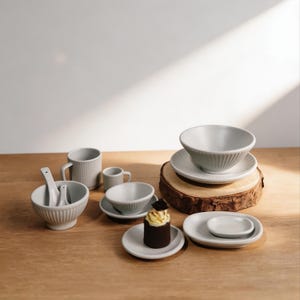 May include: A selection of light grey ceramic tableware, including bowls, plates, cups, and spoons, arranged on a wooden surface. The set includes a small cupcake with yellow icing. The pieces have a ribbed design.