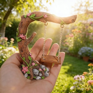 Puede incluir: Un columpio en miniatura para jardín de hadas. El columpio es de madera y cuelga de una rama marrón con flores rosas y hojas verdes. La base del columpio está hecha de rocas y más flores rosas.