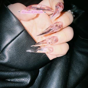 Puede incluir: Uñas postizas largas, transparentes y en forma de estilete con escamas de purpurina rosa e iridiscente incrustadas. Las uñas tienen una punta afilada y una base transparente. El texto Kawaii Kollectibles es visible en letra rosa en la imagen.
