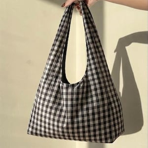 May include: A hobo-style shoulder bag featuring a black and white gingham check pattern. The bag has a wide, integrated shoulder strap and a spacious, unstructured body with a soft, fabric construction and a simple, open-top design.